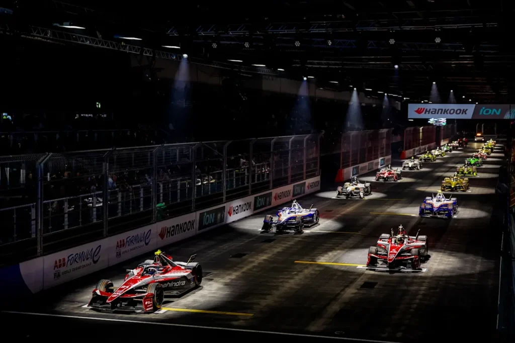 Formula E race cars lined up on a starting grid under spotlights on an indoor track section.