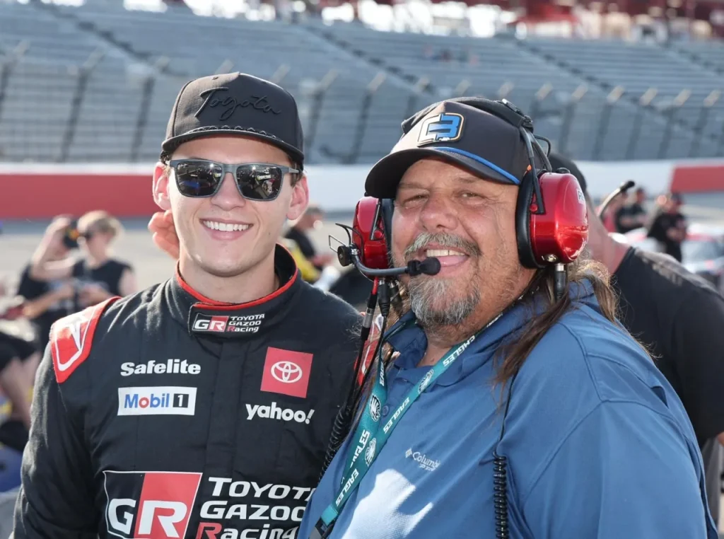 A smiling man wearing a spotter headset and cap stands beside a racing driver wearing sunglasses and a firesuit in front of track grandstands.